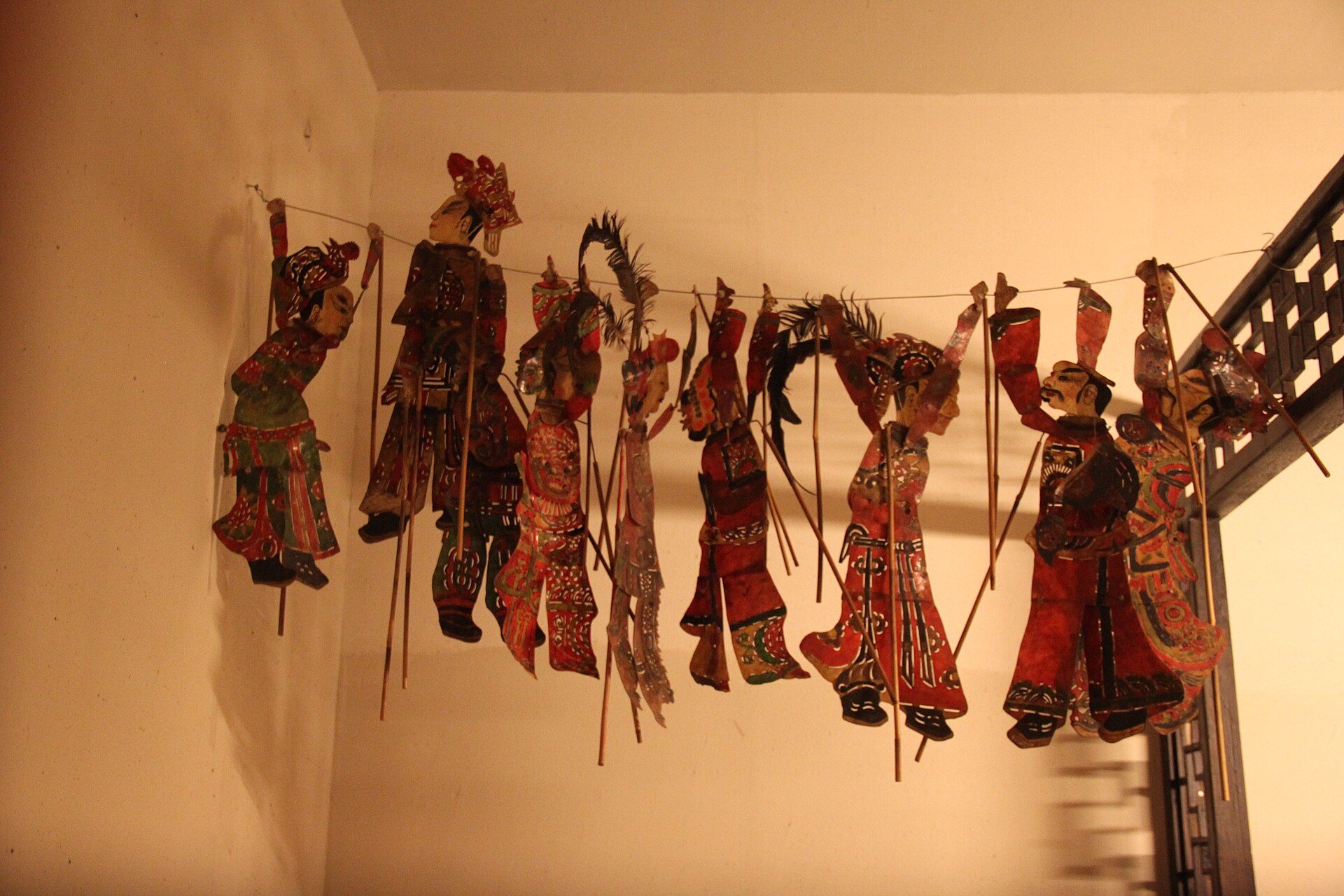 A long array of shadow-puppet figures hung before a lamp on a white wall: red and green carved hide, bamboo rods for control