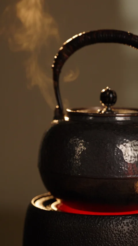 A black iron kettle heating on a charcoal stove, steam rising from the spout